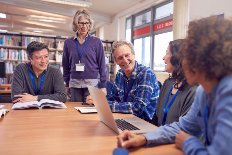 Teacher With Group Of Mature Adult Students In Class Sit Around Table And Work In College Library